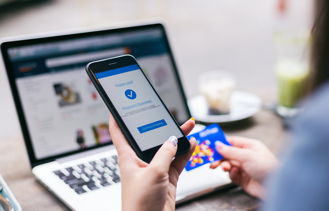 cell phone in hand to make an online purchase with a laptop behind and a woman holding a credit card